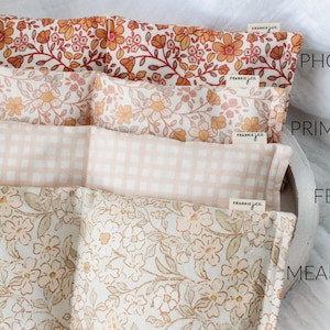 May include: Four rectangular fabric items with floral and gingham patterns. The top item is Phoebe, with orange and red flowers. Primrose has peach and yellow flowers. Fern is a peach gingham. Meadow has white and tan flowers. Each item has a Frankie & Co. tag.