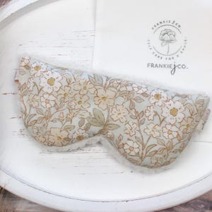 Puede incluir: M&aacute;scara para los ojos con estampado floral y un interior blanco y suave. La m&aacute;scara presenta un fondo azul claro con un patr&oacute;n de flores blancas y doradas. Un pa&ntilde;o blanco con un logotipo est&aacute; en el fondo. La m&aacute;scara est&aacute; dise&ntilde;ada para la relajaci&oacute;n y el sue&ntilde;o.