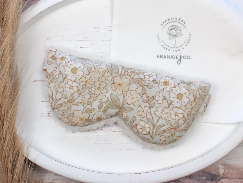 Puede incluir: M&aacute;scara para los ojos con estampado floral y un interior blanco y suave. La m&aacute;scara presenta un fondo azul claro con un patr&oacute;n de flores blancas y doradas. Un pa&ntilde;o blanco con un logotipo est&aacute; en el fondo. La m&aacute;scara est&aacute; dise&ntilde;ada para la relajaci&oacute;n y el sue&ntilde;o.