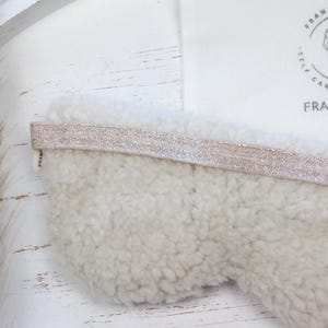 Puede incluir: Una m&aacute;scara para dormir de color crema con una superficie suave y texturizada y una banda el&aacute;stica rosa claro brillante. La m&aacute;scara se combina con una bolsa de tela blanca con el texto "FRANKIE jco." y un logotipo floral.