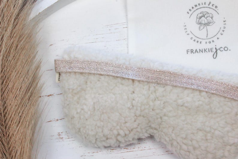 Puede incluir: Una m&aacute;scara para dormir de color crema con una superficie suave y texturizada y una banda el&aacute;stica rosa claro brillante. La m&aacute;scara se combina con una bolsa de tela blanca con el texto "FRANKIE jco." y un logotipo floral.