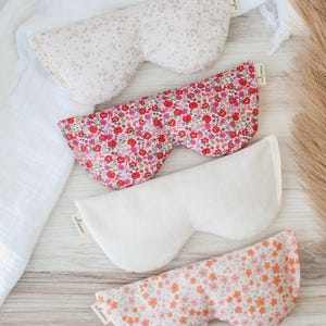 May include: Four eye masks with floral and dotted patterns. The masks are in shades of pink, white, and peach. Each mask has a small tag on the side. The masks are arranged on a light wooden surface.