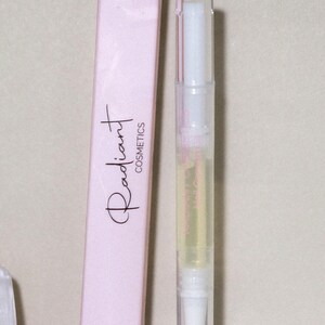 May include: A light pink box and a clear nail oil pen are displayed. The box has the words "Radiant Cosmetics" in a script font. The pen is filled with a yellow liquid and has a brush tip. The box reads "Nail Growth Oil NET WT. 10 oz/3 ml."