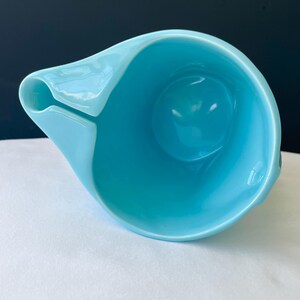 Pode incluir: Um jarro de cer&acirc;mica azul claro com um design assim&eacute;trico &uacute;nico. O jarro apresenta um bico e uma al&ccedil;a, e um interior arredondado. O acabamento brilhante reflete a luz, destacando as curvas suaves do recipiente. O jarro est&aacute; sobre uma superf&iacute;cie branca.