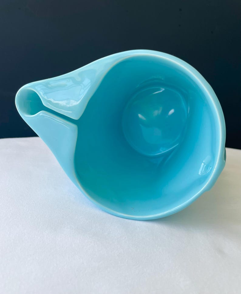 Pode incluir: Um jarro de cer&acirc;mica azul claro com um design assim&eacute;trico &uacute;nico. O jarro apresenta um bico e uma al&ccedil;a, e um interior arredondado. O acabamento brilhante reflete a luz, destacando as curvas suaves do recipiente. O jarro est&aacute; sobre uma superf&iacute;cie branca.