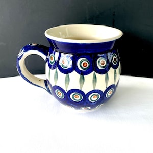 May include: A ceramic mug with a blue and white floral pattern. The mug has a rounded body, a handle, and a wide rim. The design includes blue circles with red and green accents, and green leaf shapes. The interior is cream-colored.