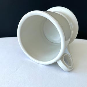 Op de afbeelding: Een witte keramische mok met een uniek ontwerp. De mok heeft een brede, cilindrische body en een handvat met een cirkelvormige opening. De binnen- en buitenkant zijn effen wit, met een licht off-white accent op het handvat.