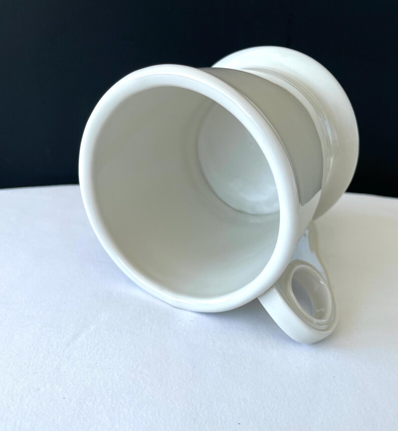 Op de afbeelding: Een witte keramische mok met een uniek ontwerp. De mok heeft een brede, cilindrische body en een handvat met een cirkelvormige opening. De binnen- en buitenkant zijn effen wit, met een licht off-white accent op het handvat.