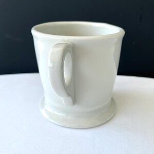 Op de afbeelding: Een witte keramische mok met een afgeronde handgreep en een brede basis. De mok heeft een eenvoudig, klassiek ontwerp en is geschikt voor warme dranken. De achtergrond is contrasterend zwart en wit.
