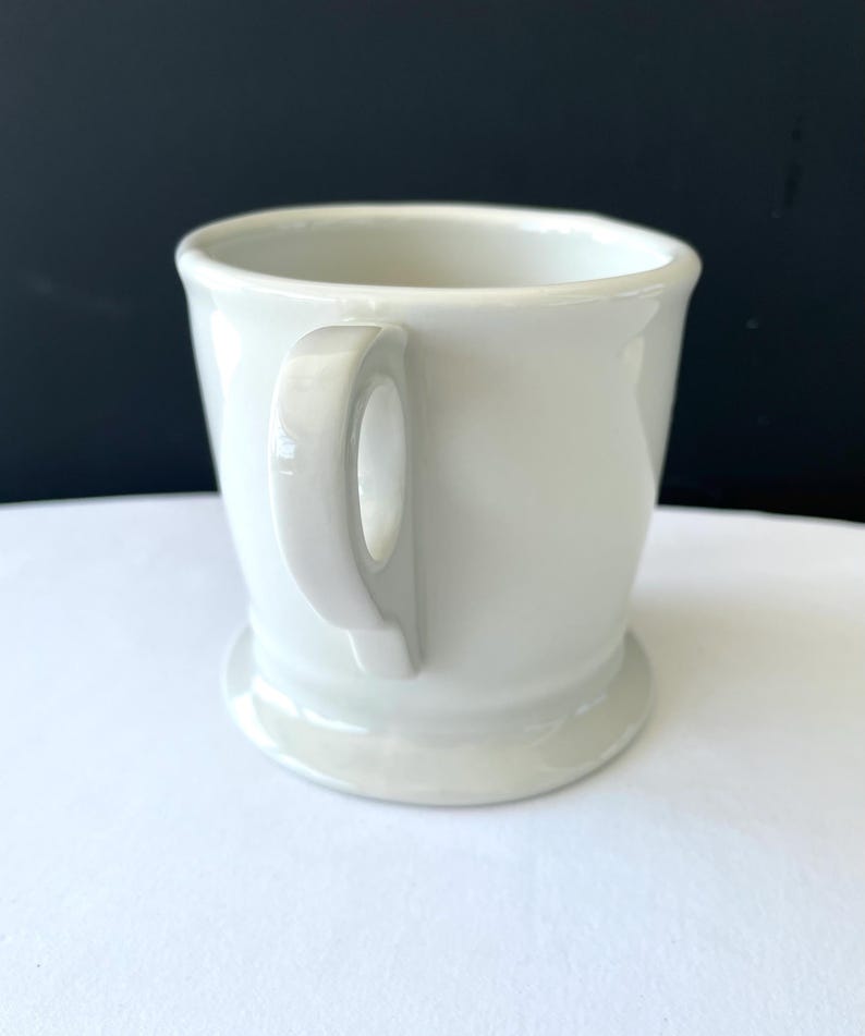 Op de afbeelding: Een witte keramische mok met een afgeronde handgreep en een brede basis. De mok heeft een eenvoudig, klassiek ontwerp en is geschikt voor warme dranken. De achtergrond is contrasterend zwart en wit.