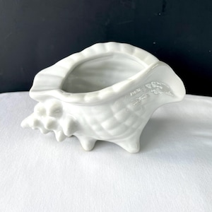May include: A white ceramic seashell-shaped planter. The planter has a textured surface and a wide opening. The shell is supported by three small feet. The overall design is a detailed and realistic representation of a seashell.