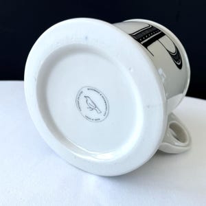 Op de afbeelding: Witte keramische mok met een zwart letterontwerp. De onderkant van de mok heeft een ronde stempel met een vogelillustratie en de tekst "MICROWAVE & DISHWASHER SAFE" en "MADE IN CHINA". De mok heeft een gebogen handvat.