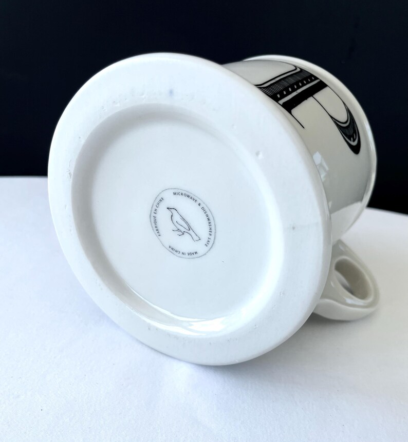 Op de afbeelding: Witte keramische mok met een zwart letterontwerp. De onderkant van de mok heeft een ronde stempel met een vogelillustratie en de tekst "MICROWAVE & DISHWASHER SAFE" en "MADE IN CHINA". De mok heeft een gebogen handvat.