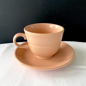 May include: A solid light peach-colored ceramic teacup and matching saucer set. The teacup features a rounded handle and a series of horizontal embossed rings around the exterior. The saucer has a matching circular design with concentric rings.