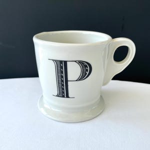 Op de afbeelding: Witte keramische mok met een zwarte letter "P" in een decoratief lettertype. De mok heeft een afgeronde basis en een gebogen handvat. De mok staat op een wit oppervlak met een zwarte achtergrond. De mok is ongeveer 10 cm hoog.