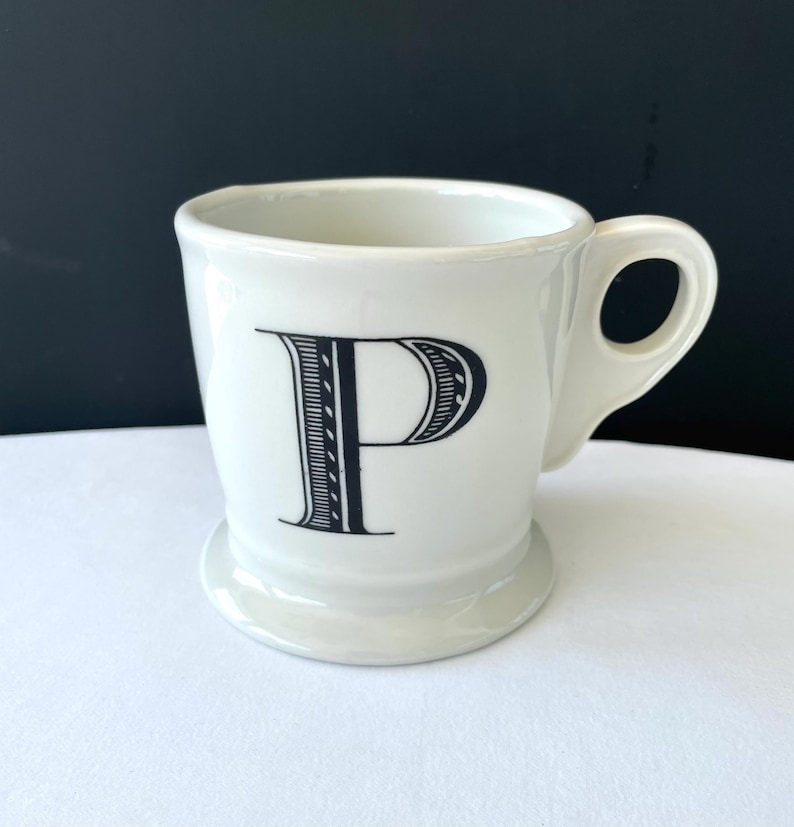 Op de afbeelding: Witte keramische mok met een zwarte letter "P" in een decoratief lettertype. De mok heeft een afgeronde basis en een gebogen handvat. De mok staat op een wit oppervlak met een zwarte achtergrond. De mok is ongeveer 10 cm hoog.