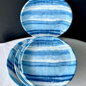May include: Set of four round ceramic plates with a blue and white striped design. The plates have a glossy finish and are arranged in a stack, showcasing the watercolor-like pattern. The plates are suitable for serving food.