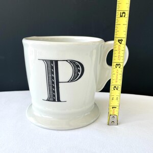 Op de afbeelding: Een witte keramische mok met de zwarte letter "P" op de voorkant. De mok heeft een handvat en een kleine voet. De mok is ongeveer 10 cm hoog.