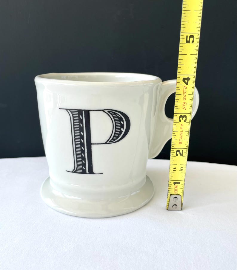 Op de afbeelding: Een witte keramische mok met de zwarte letter "P" op de voorkant. De mok heeft een handvat en een kleine voet. De mok is ongeveer 10 cm hoog.