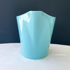 Pode incluir: Um jarro de cer&acirc;mica azul claro com uma borda ondulada &uacute;nica. O jarro tem um acabamento liso e brilhante e parece estar vazio. Texto pequeno e um gr&aacute;fico vermelho s&atilde;o vis&iacute;veis na lateral. O jarro est&aacute; sobre um fundo azul escuro.