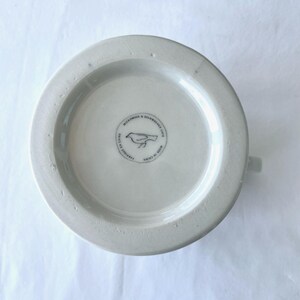 Op de afbeelding: Een witte keramische mok, van onderaf gezien, met een klein handvat. De basis heeft een cirkelvormig embleem met een vogelillustratie en de tekst "MICROWAVE & DISHWASHER SAFE" en "FABRIQUE EN CHINE".