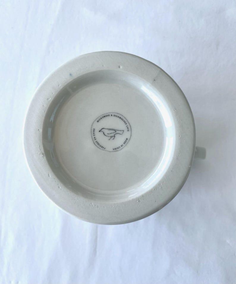 Op de afbeelding: Een witte keramische mok, van onderaf gezien, met een klein handvat. De basis heeft een cirkelvormig embleem met een vogelillustratie en de tekst "MICROWAVE & DISHWASHER SAFE" en "FABRIQUE EN CHINE".