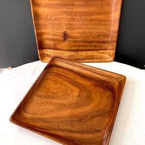 May include: Two square wooden plates with rounded corners and a smooth, polished finish. The plates feature a rich, warm brown color with visible natural wood grain patterns throughout the surface, showcasing a simple and functional design.