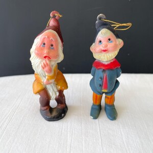 May include: Two vintage gnome figurines. One gnome wears a brown hat and yellow coat, with a white beard and a sad expression. The other gnome has a blue coat, red collar, and a black hat. Both have gold string for hanging.