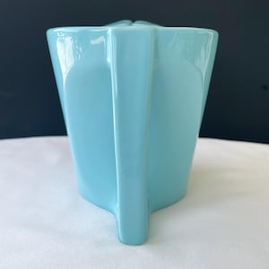 Pode incluir: Um jarro de cer&acirc;mica azul claro com um design geom&eacute;trico. O jarro tem uma al&ccedil;a e um bico. O jarro est&aacute; sobre uma superf&iacute;cie branca com um fundo escuro.