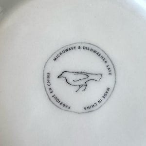 Op de afbeelding: Close-up van een wit keramisch bord met een cirkelvormige stempel. De stempel toont een vogelillustratie en de tekst "MICROWAVE & DISHWASHER SAFE", "FABRIQUE EN CHINE" en "MADE IN CHINA".
