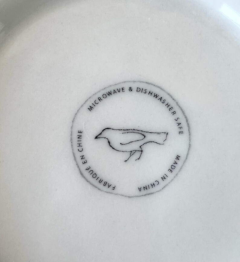 Op de afbeelding: Close-up van een wit keramisch bord met een cirkelvormige stempel. De stempel toont een vogelillustratie en de tekst "MICROWAVE & DISHWASHER SAFE", "FABRIQUE EN CHINE" en "MADE IN CHINA".