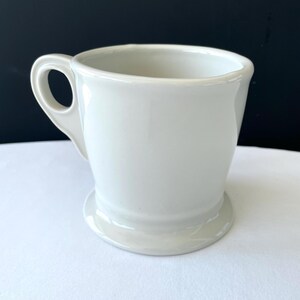 Op de afbeelding: Een witte keramische mok met een afgeronde basis en een gebogen handvat. De mok heeft een gladde, glanzende afwerking en een eenvoudig, klassiek ontwerp. De mok is leeg en klaar om gevuld te worden met een drankje.