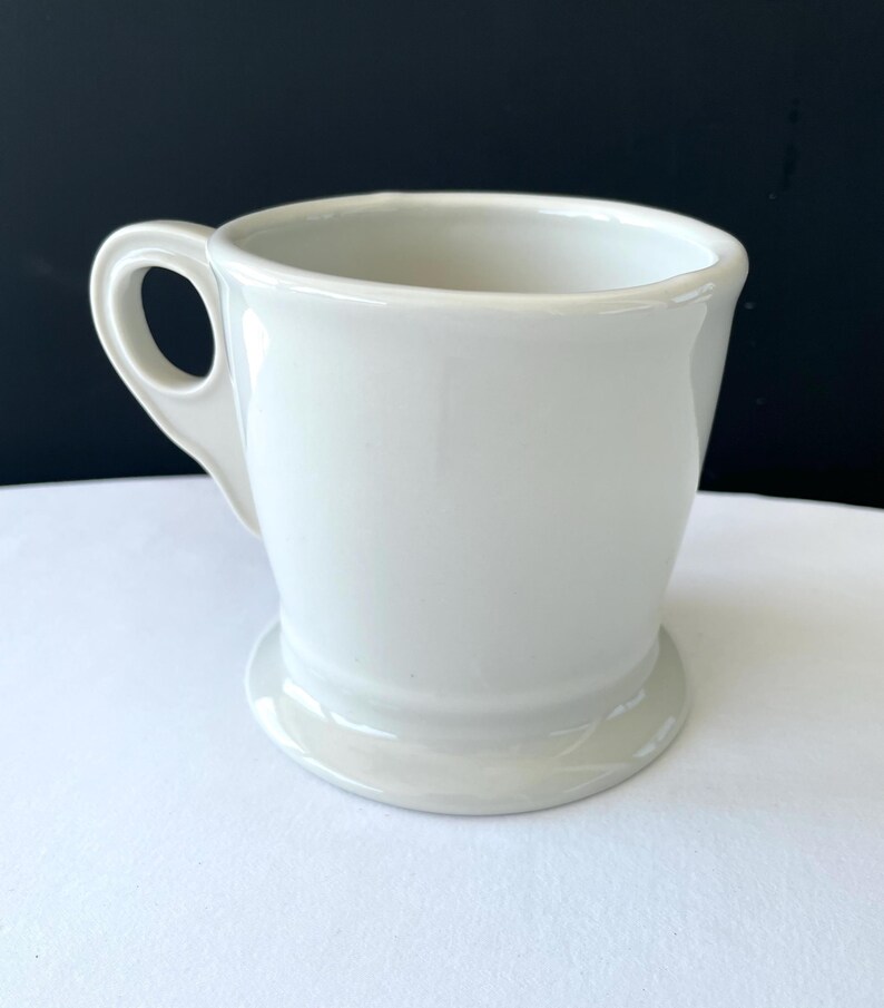 Op de afbeelding: Een witte keramische mok met een afgeronde basis en een gebogen handvat. De mok heeft een gladde, glanzende afwerking en een eenvoudig, klassiek ontwerp. De mok is leeg en klaar om gevuld te worden met een drankje.