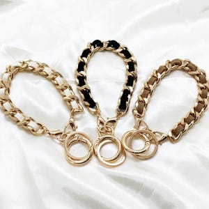 May include: Three gold chain keychains with different colored fabric accents. The chains are linked together with a gold ring. The fabric accents are black, white, and brown.