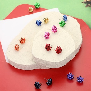 May include: Assortment of colourful bow-shaped earrings displayed on a white surface. The earrings are made of metal and have a shiny finish. The colours include red, green, blue, purple, gold, and silver.