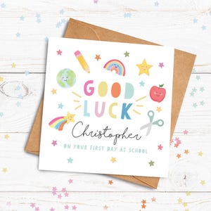 May include: A square greeting card with a white background featuring the text GOOD LUCK in colorful letters, the name Christopher in black script, and the text ON YOUR FIRST DAY AT SCHOOL. Illustrations include a pencil, rainbow, star, globe, apple, and scissors.