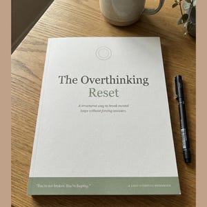 May include: A workbook titled "The Overthinking Reset" with the text "A structured way to break mental loops without forcing answers." A black pen rests on the book's right side. A mug is in the background.