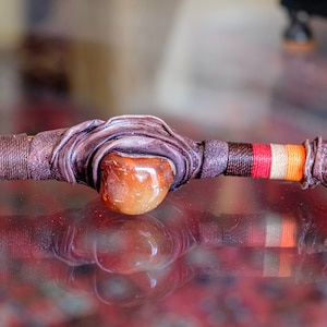 May include: A brown leather wrapped wand with a red, white, and orange striped band and a large amber gemstone in the center.