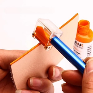 May include: A leather edge painting tool with a blue metal handle and a clear plastic head containing an orange applicator wheel, shown applying orange paint to the edge of a piece of light tan leather next to a bottle of orange paint.