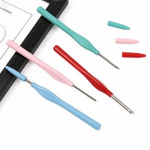 May include: A set of four crochet hooks with ergonomic handles in light blue, light pink, red, and teal. Each hook features a metallic tip and a matching removable protective cap. The tools are arranged diagonally alongside a black picture frame.