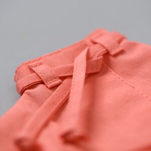 May include: Close-up of a coral pink fabric with a tied waistband. The fabric is soft and has a subtle texture.