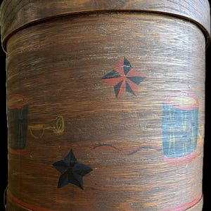 Op de afbeelding: Een ronde houten opbergdoos met een rood, zwart en gouden geschilderd ontwerp. Het ontwerp bevat sterren, een trompet en een trommel. De doos is gemaakt van hout en heeft een natuurlijke houten afwerking.