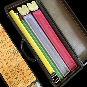 K&ouml;nnte beinhalten: Ein offenes Mahjong-Set im Vintage-Stil in einem dunkelbraunen Koffer. Im Inneren befinden sich farbige Gestelle in Gelb, Gr&uuml;n, Klar und Pink. Das Set enth&auml;lt beige Spielsteine mit chinesischen Schriftzeichen und Symbolen.