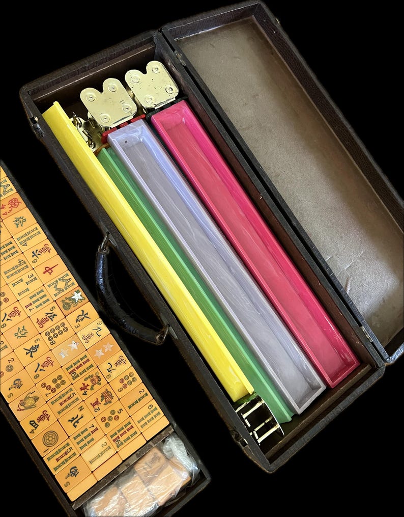 K&ouml;nnte beinhalten: Ein offenes Mahjong-Set im Vintage-Stil in einem dunkelbraunen Koffer. Im Inneren befinden sich farbige Gestelle in Gelb, Gr&uuml;n, Klar und Pink. Das Set enth&auml;lt beige Spielsteine mit chinesischen Schriftzeichen und Symbolen.