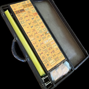 K&ouml;nnte beinhalten: Ein Mahjong-Set in einem offenen schwarzen Koffer. Das Set enth&auml;lt zahlreiche rechteckige Spielsteine mit Symbolen und Zahlen, einen langen gelben Stab und ein in Kunststoff verpacktes Paket. Der Koffer hat einen Griff und Metallverschl&uuml;sse.