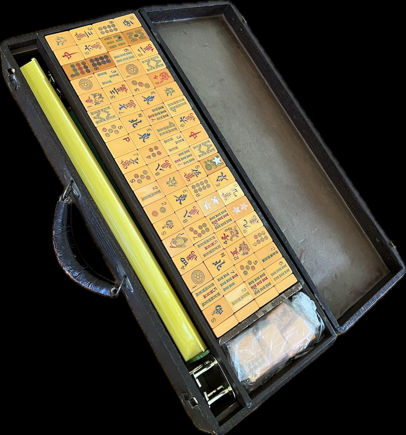 K&ouml;nnte beinhalten: Ein Mahjong-Set in einem offenen schwarzen Koffer. Das Set enth&auml;lt zahlreiche rechteckige Spielsteine mit Symbolen und Zahlen, einen langen gelben Stab und ein in Kunststoff verpacktes Paket. Der Koffer hat einen Griff und Metallverschl&uuml;sse.
