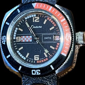 May include: A vintage Endura watch with a black dial, white hour markers, and a date display at the 3 o'clock position. The watch has a black bezel with orange accents and a black leather strap. The watch face displays the text "Endura".