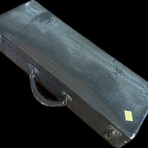 K&ouml;nnte beinhalten: Ein rechteckiger, schwarzer Vintage-Koffer mit strukturierter Oberfl&auml;che und gebogenem Griff. Der Koffer hat Metallverschl&uuml;sse und Scharniere, und ein kleiner gelber Aufkleber ist an der Seite angebracht. Der Koffer scheint aus einem strapazierf&auml;higen Material zu bestehen.