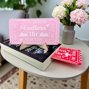 May include: A pink decorative ticket with the text "Endless TBR Book Club" is propped up on a book. A red ticket with the text "Fire Seeker" is used as a bookmark. A vase of pink and white flowers sits nearby.