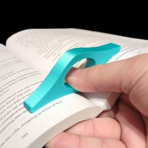 May include: A turquoise blue plastic page holder for books. The page holder is shaped like a curved triangle with a hole in the center for a finger to go through. The page holder is being used to hold a book open.